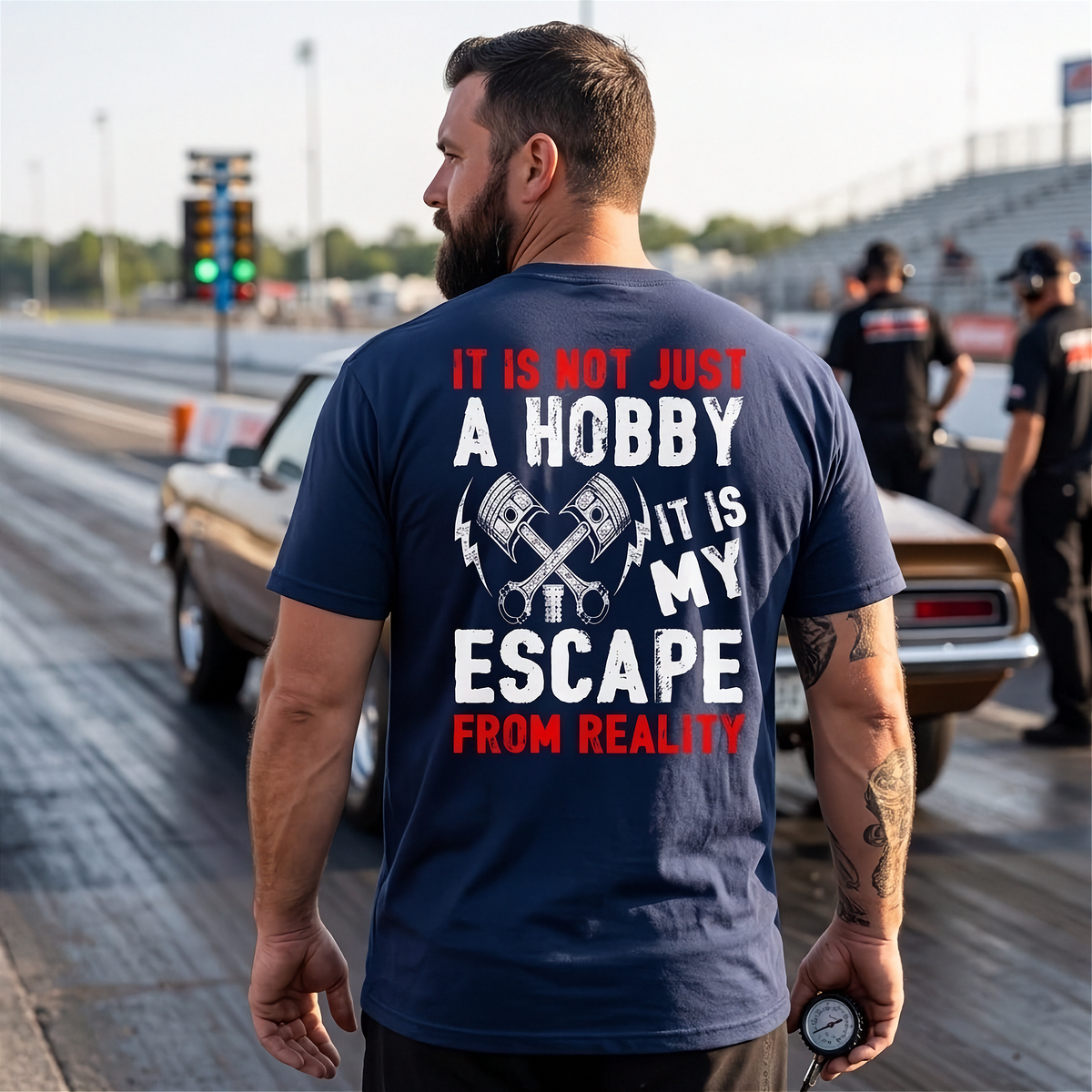 A man standing on a drag strip wearing a navy blue t-shirt with a graphic print on the back. The print features crossed pistons and the text 'IT IS NOT JUST A HOBBY IT IS MY ESCAPE FROM REALITY' in white and red.