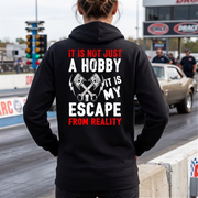 A person standing on a drag strip wearing a black hoodie from the back, which features a graphic print in white and red that reads 'IT IS NOT JUST A HOBBY IT IS MY ESCAPE FROM REALITY' with crossed pistons. In the background, a classic car is visible on the track.