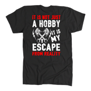 A black short-sleeve t-shirt with a graphic print on the front. The design features crossed pistons and bolts with the text 'IT IS NOT JUST A HOBBY IT IS MY ESCAPE FROM REALITY' in distressed white and red lettering, set against a plain white background.