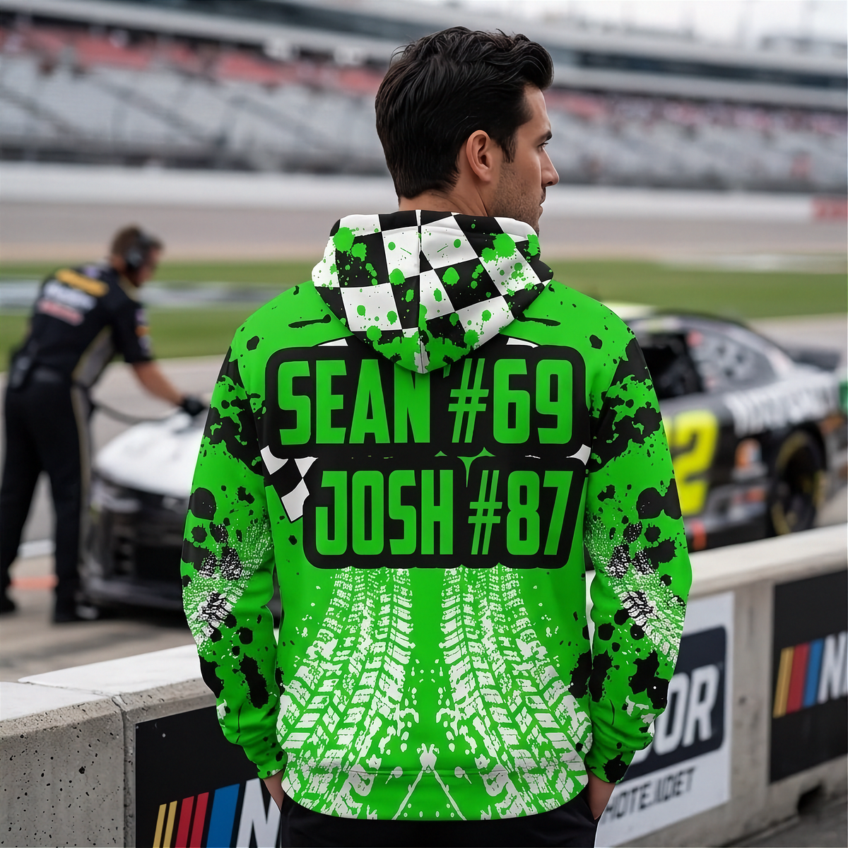 A man stands at a race track barrier wearing a bright neon green and black pullover hoodie with a checkered hood, tire-track print, and large text reading 'SEAN #69 JOSH #87' on the back. A race car and crew are blurred in the background.