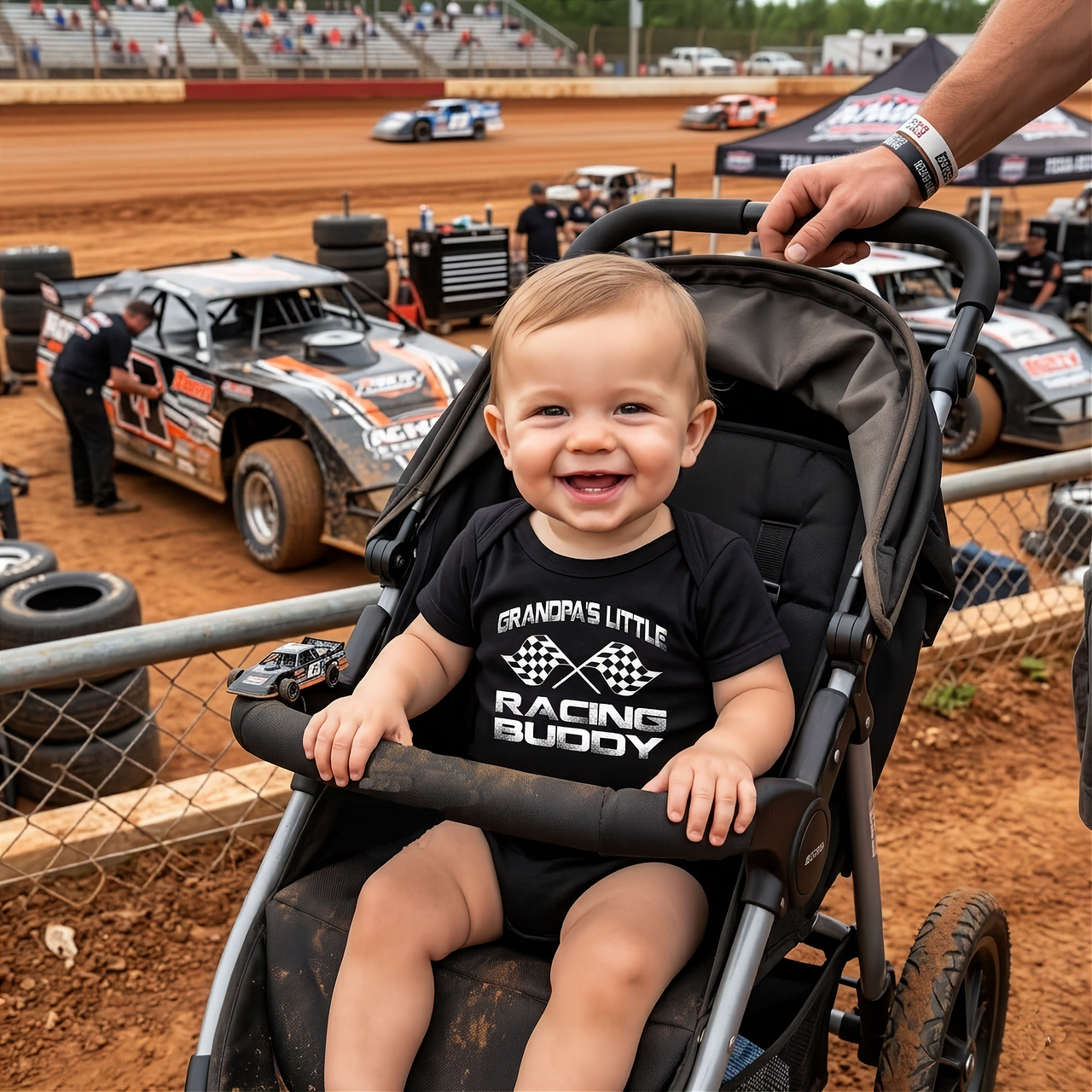 Child in a stroller at a dirt track with racing cars in the background
