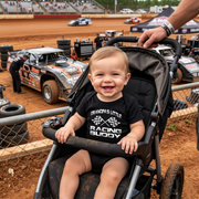 Child in a stroller at a dirt track with racing cars in the background