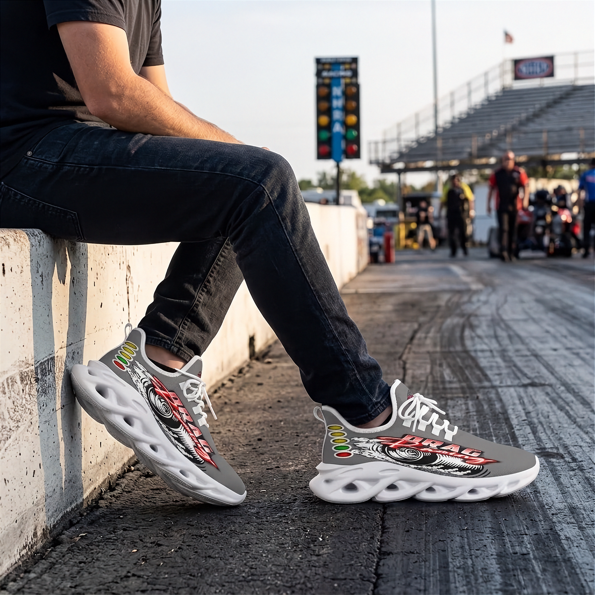 A person sitting on a concrete barrier at a drag strip wearing gray and white graphic sneakers with red, black, and yellow details. The background shows a racetrack, bleachers, and other people in the distance.