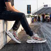 A person sitting on a concrete barrier at a drag strip wearing gray and white graphic sneakers with red, black, and yellow details. The background shows a racetrack, bleachers, and other people in the distance.