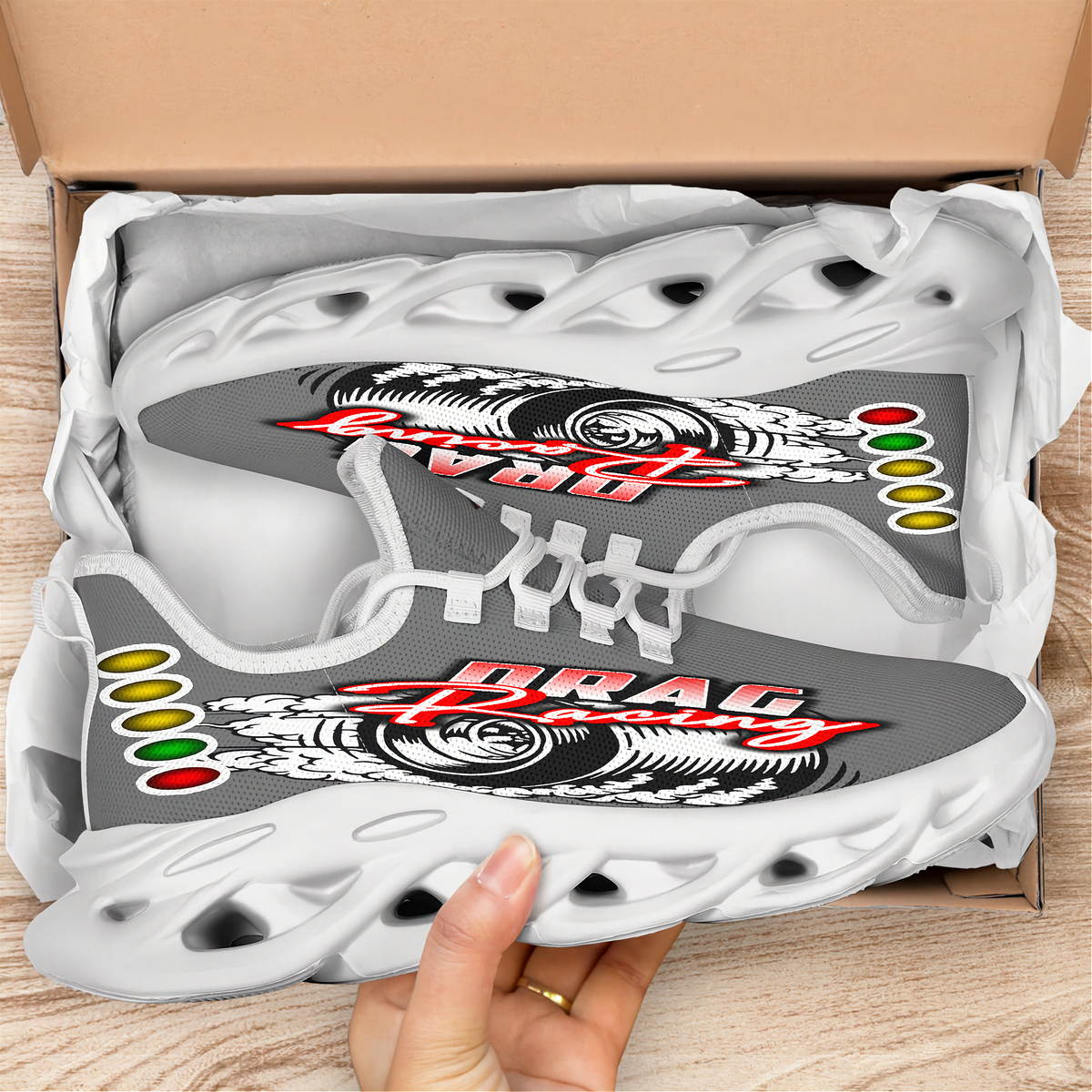 A pair of custom gray and white sneakers with a 'DRAC' graphic and Rastafarian flag accents, shown inside an open cardboard shoebox with a hand holding the front shoe.