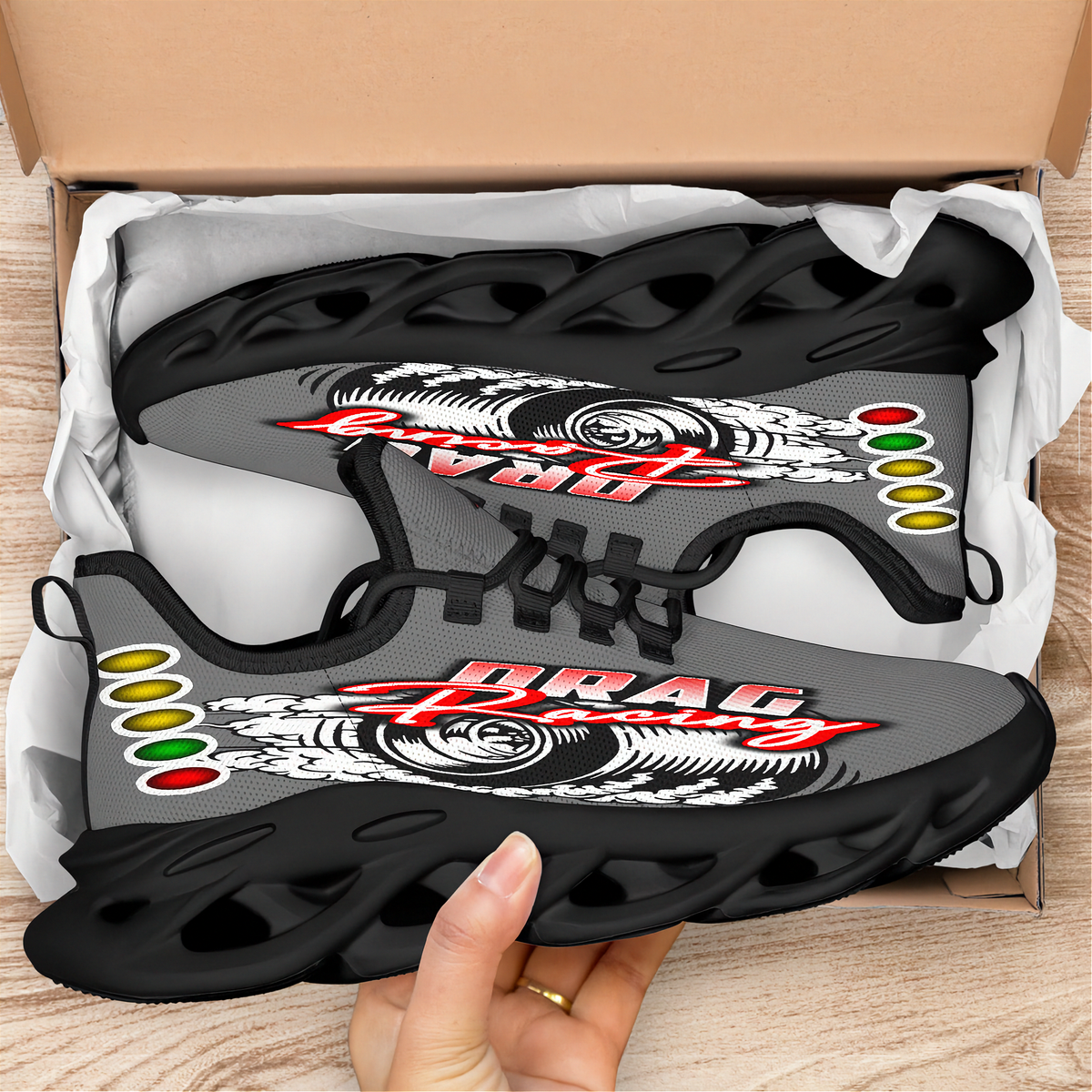 A pair of custom black and gray performance sneakers with a white and red 'DRAG RACING' graphic and Rastafarian flag accents, shown inside an open cardboard box with a hand holding one shoe.