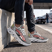 A person sitting on a concrete barrier at a racetrack wearing gray and white graphic sneakers with red, yellow, and green accents and a black-and-white wing design. A car is visible in the background under a canopy.