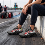 A person sitting on a concrete barrier at a racetrack wearing black and gray Drad sneakers with a red, white, and green graphic design. The background shows a blurred racetrack scene with tires and track crew.