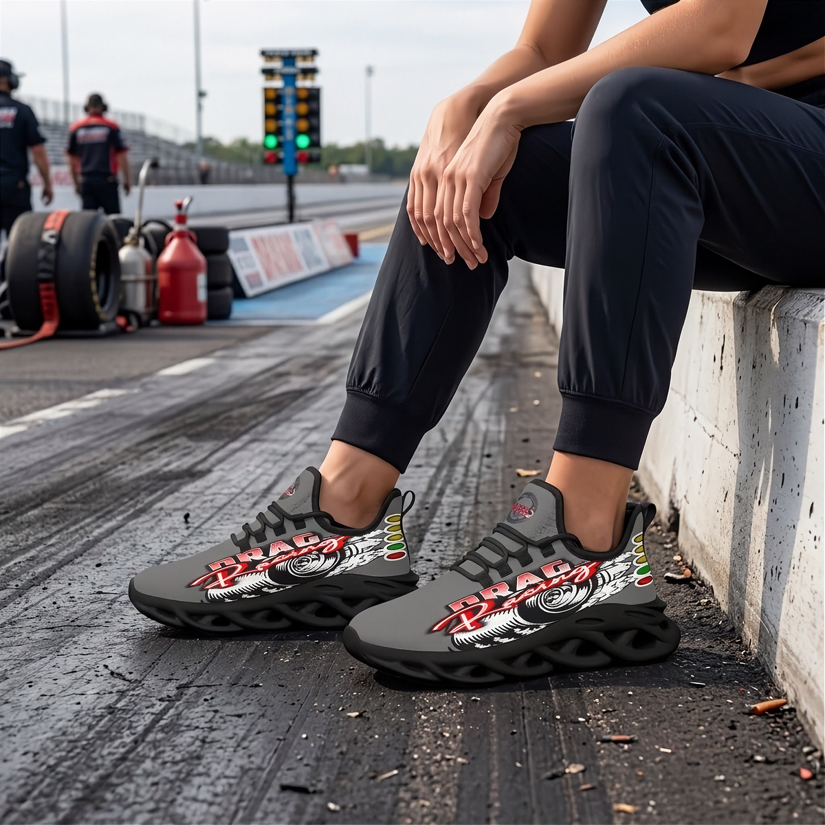 A person sitting on a concrete barrier at a racetrack wearing black and gray Drad sneakers with a red, white, and green graphic design. The background shows a blurred racetrack scene with tires and track crew.