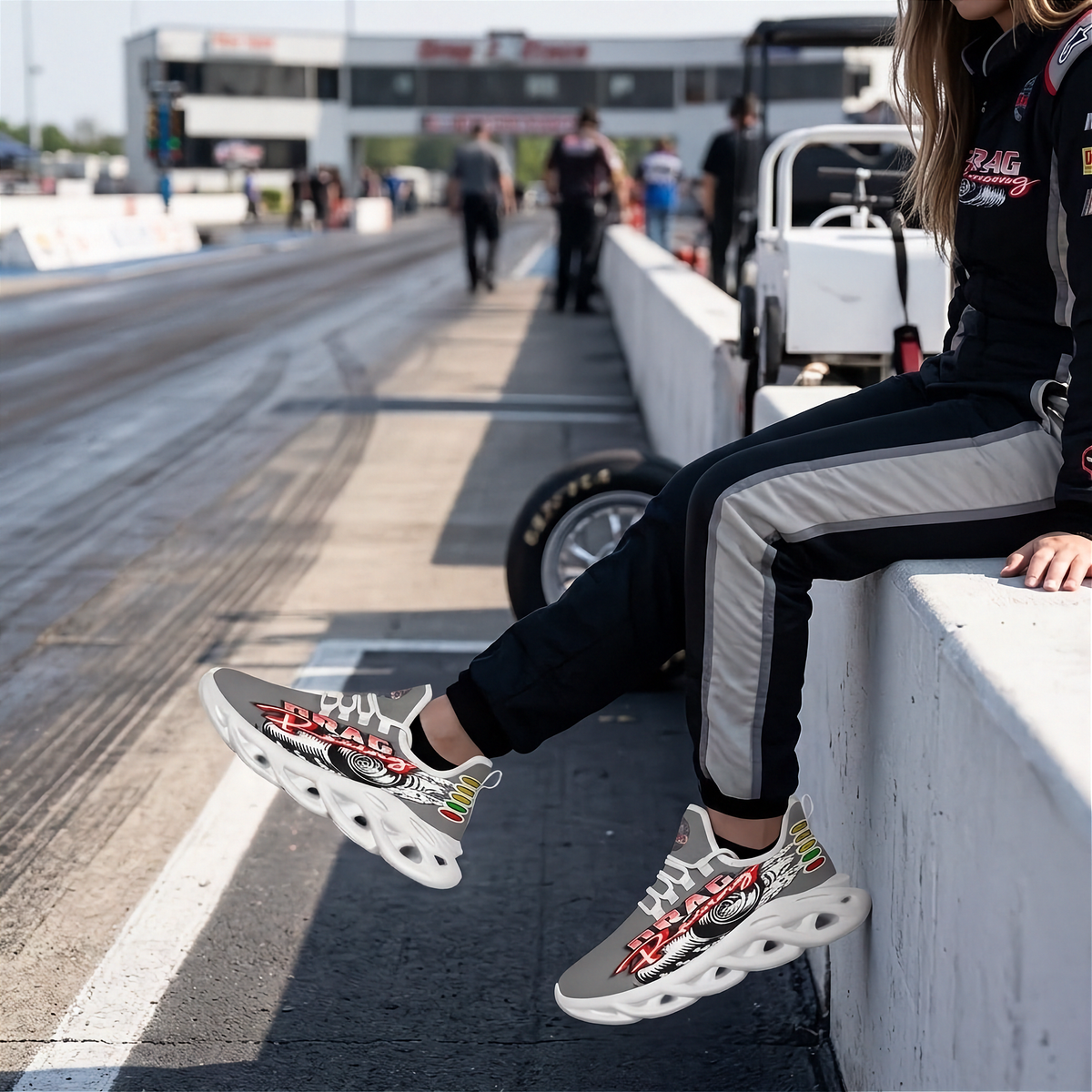 A person sitting on a concrete barrier at a racetrack wearing gray and black drag racing track pants and gray chunky sneakers with red, white, and black graphics.