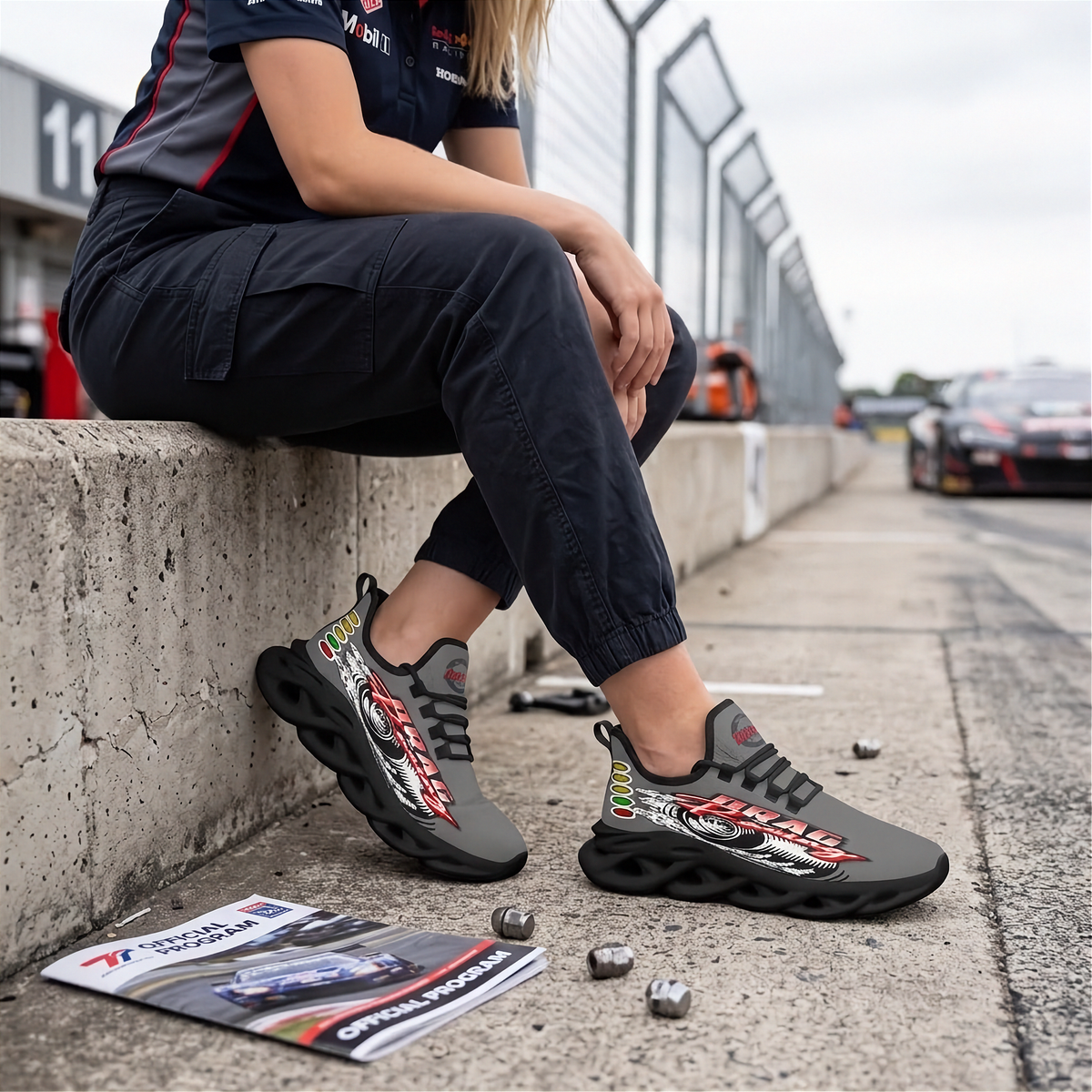 A person sitting on a concrete barrier at a racetrack wearing dark gray and black racing sneakers with red, white, and yellow graphic accents. The person is also wearing a dark navy team shirt and cargo pants.