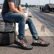 A man sitting on stacked tires at a racetrack wearing dark gray and black graphic sneakers with red, white, and green accents. The background shows a blurred racetrack with other people and equipment.