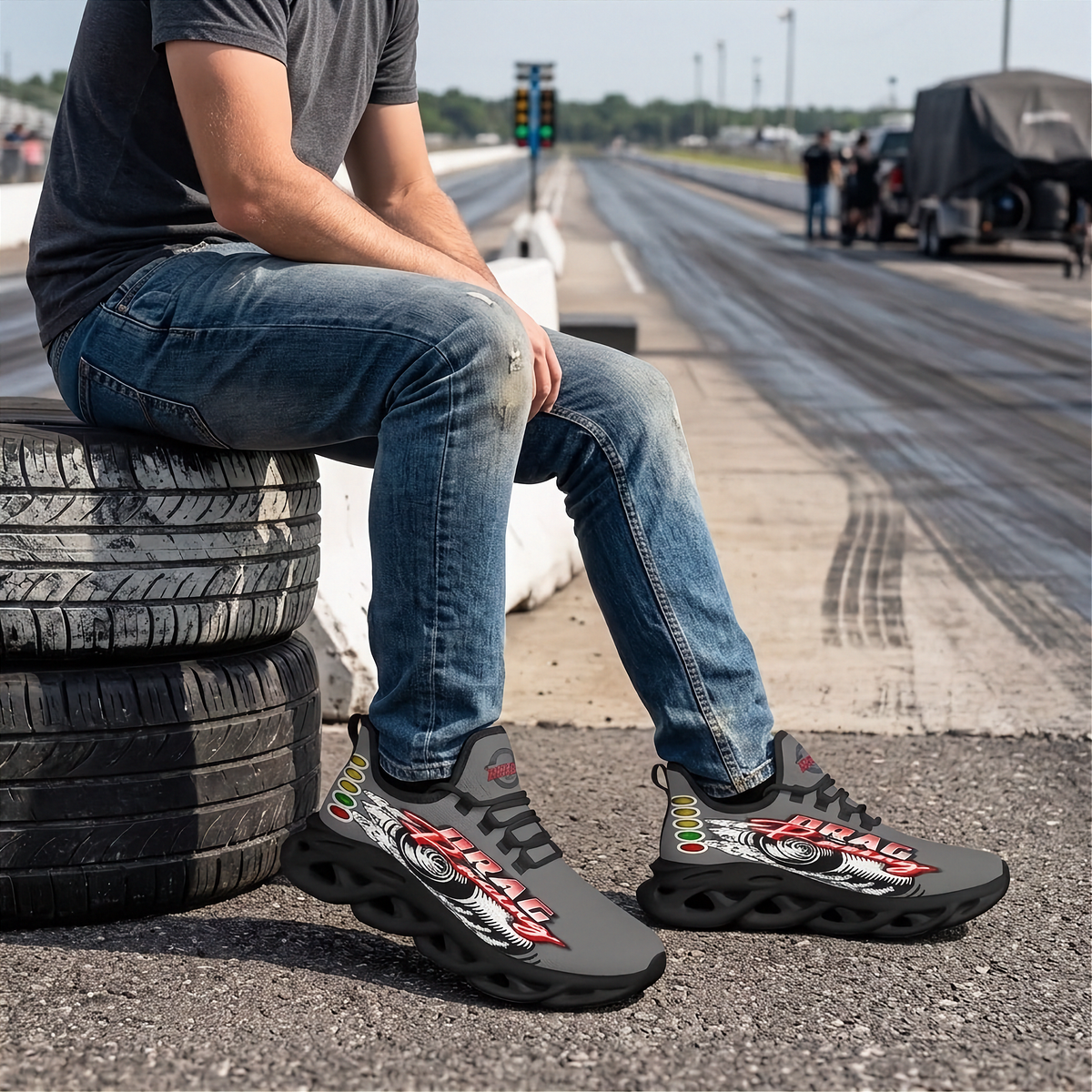 A man sitting on stacked tires at a racetrack wearing dark gray and black graphic sneakers with red, white, and green accents. The background shows a blurred racetrack with other people and equipment.