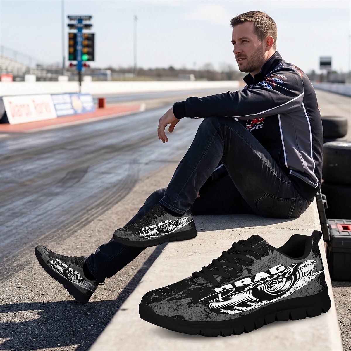 A man sits on a concrete barrier at a drag strip wearing black and gray drag-racing team apparel, with a matching pair of black and gray graphic drag shoes displayed in the foreground. The shoes feature a white tiger graphic and the text 'DRAG RACING'.