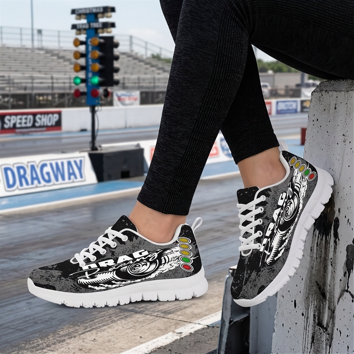 A person wearing black ribbed leggings and black-and-white graphic sneakers with white laces, sitting on a concrete track barrier with a drag strip and stadium in the background.