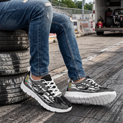 A person wearing distressed blue jeans and black-and-white graphic sneakers sits on stacked tires in a garage or workshop setting. The shoes feature white laces, thick white soles, and a black upper with white abstract line art and small red, green, and yellow accents.