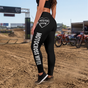 Black high-waisted motocross leggings with white 'MOTOCROSS WIFE' text and a heart graphic on the back, modeled on a dirt track with motocross bikes in the background.