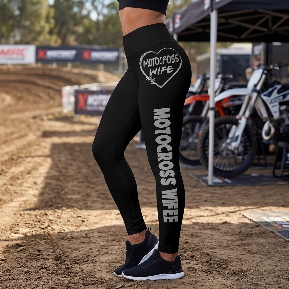 Black athletic leggings worn by a model on a dirt track, featuring 'MOTOCROSS WIFE' printed in white inside a heart on the upper thigh and vertically down the left leg.