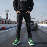 Man wearing a black hoodie and green sneakers on a racetrack with a car in the background.
