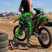 A woman wearing bright green, black, and white graphic motocross leggings stands next to a green dirt bike on a dirt track, viewed from behind.