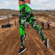 A person wearing high-waisted athletic leggings with a bold green, black, and gray motocross helmet graphic, standing on a dirt track with racers in the background.