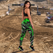 A woman wearing high-waisted athletic leggings with a green, black, and gray abstract pattern, a black sports bra, and black sneakers, standing on a dirt track with motocross bikes in the background.