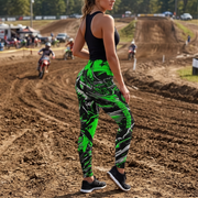 A woman stands on a dirt motocross track wearing bright neon green and black abstract-print leggings, a black tank top, and black sneakers, with riders and track terrain blurred in the background.