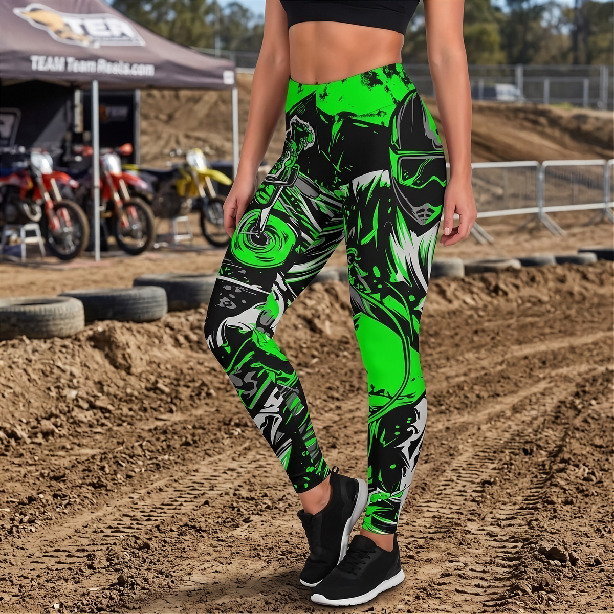 A person standing on a dirt motocross track wearing high-waisted athletic leggings with a bold black, neon green, and white abstract graphic print. The background shows a dirt track, tires, and a canopy tent with motorcycles.