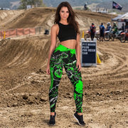 A woman stands on a dirt motocross track wearing high-waisted athletic leggings with a black and neon green graphic print, a black crop top, and black sneakers.