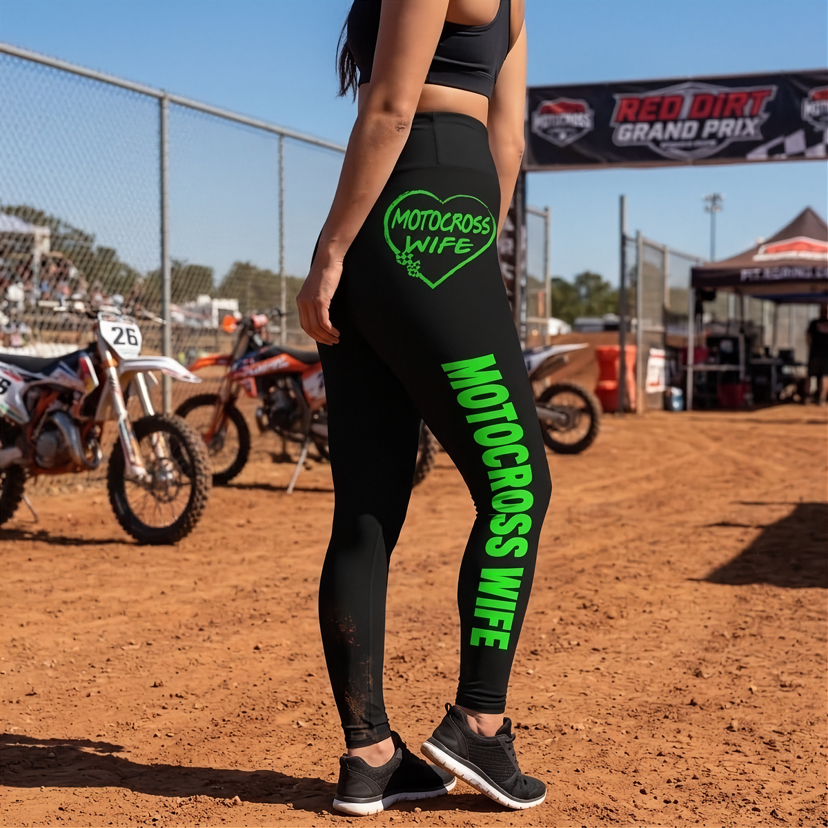 A woman wearing black leggings with bright green 'MOTOCROSS WIFE' text on the leg and a matching heart graphic on the hip, standing on a dirt track with motocross bikes and a fence in the background.