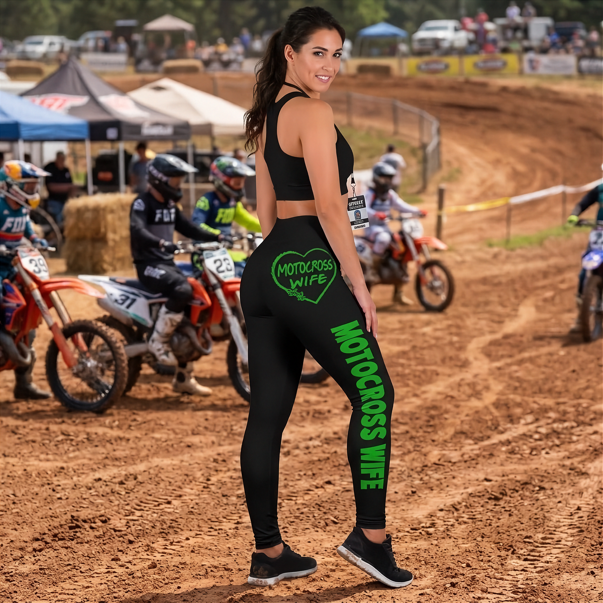 A woman stands on a dirt motocross track wearing black athletic leggings with bright green 'MOTOCROSS WIFE' text and a green heart graphic on the back, alongside a matching black sports bra. Dirt bikes and event tents are visible in the background.