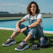 Woman sitting on grass by a pool wearing checkered sneakers and overalls, with a helmet nearby.