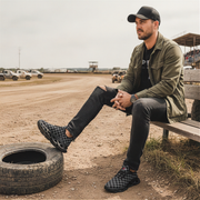 Man sitting on a tire next to a dirt track wearing a green jacket and grey checkered shoes and black cap.