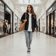 Woman walking in a shopping mall wearing a black jacket, white shirt, ripped jeans, and sneakers.