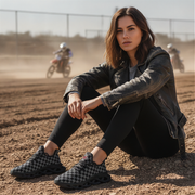 Woman sitting on a dirt track wearing checkered shoes, with motorcycles in the background.