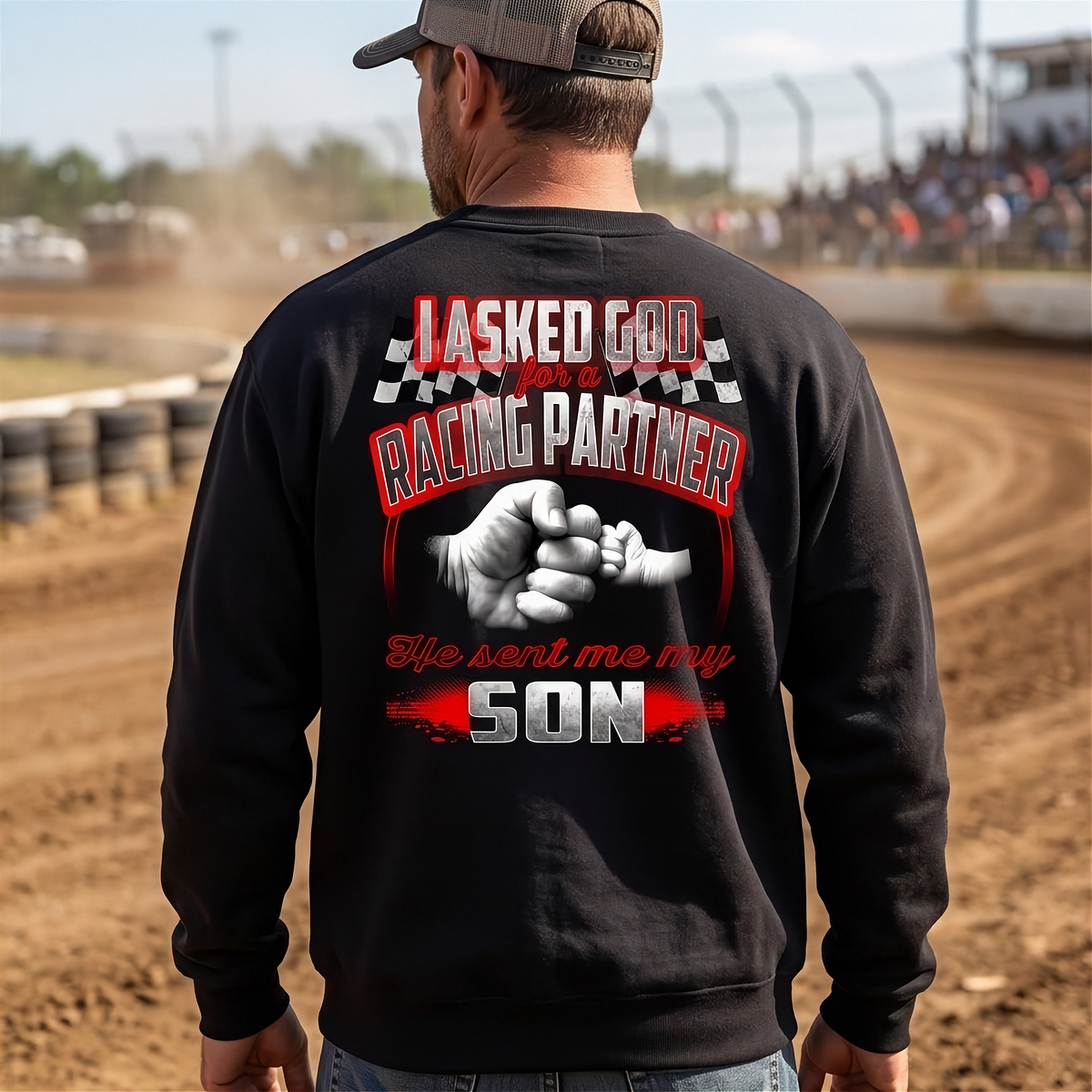 A man viewed from behind wearing a black crewneck sweatshirt with a large racing-themed graphic on the back. The graphic features the text 'I ASKED GOD FOR A RACING PARTNER' and 'He sent me my SON' in red, white, and black, along with a fist-bump illustration.