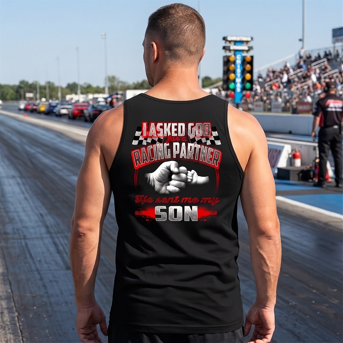 A man standing on a drag racing track, viewed from behind, wearing a black tank top with a large graphic print on the back. The graphic features a fist bump illustration and the text 'I ASKED GOD FOR RACING PARTNER' and 'She made me my SON' in red and white lettering.