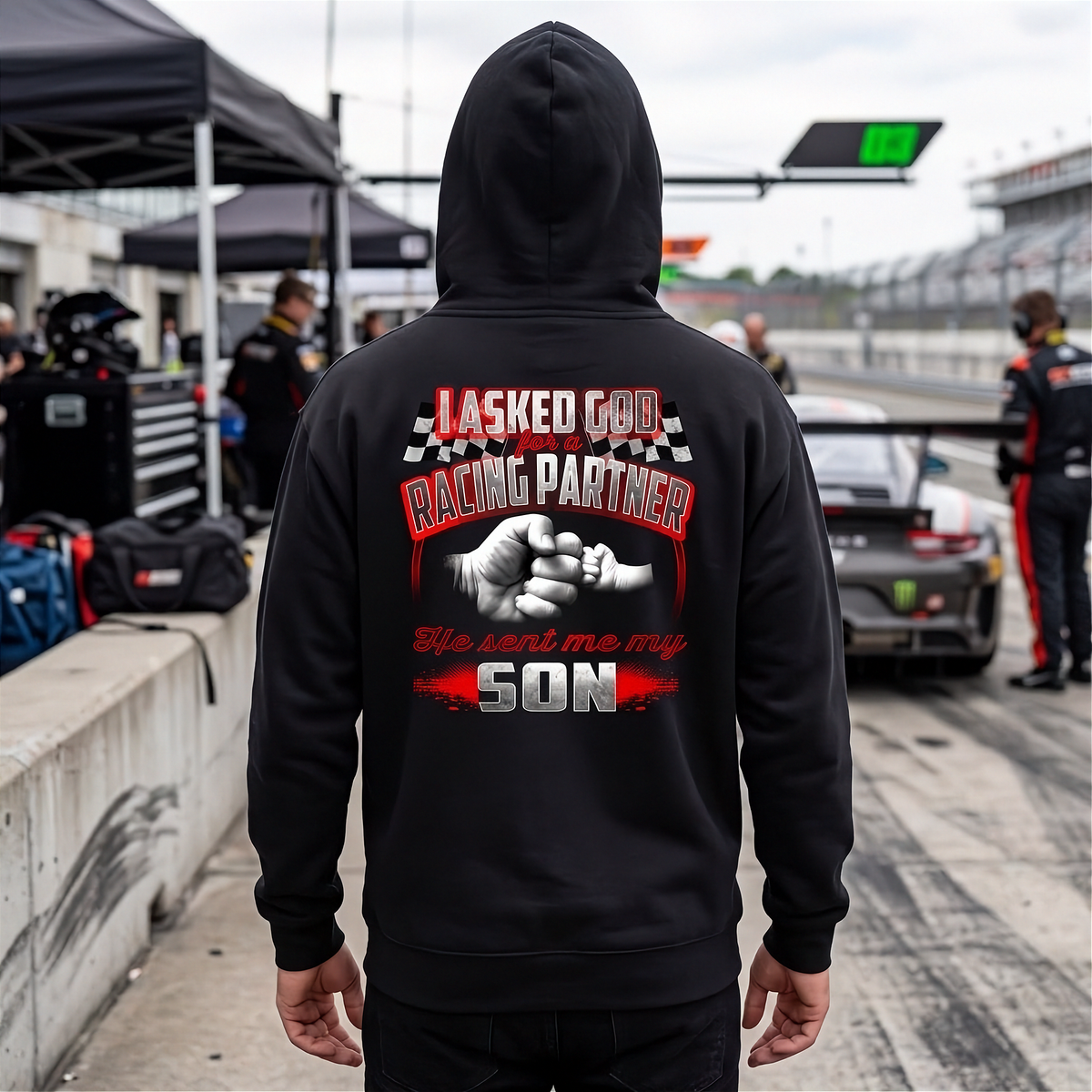 A person standing on a racetrack wearing a black hoodie from the back, which features a large graphic print with the text 'I ASKED GOD FOR A RACING PARTNER He sent me my SON' and a fist-bump design. 