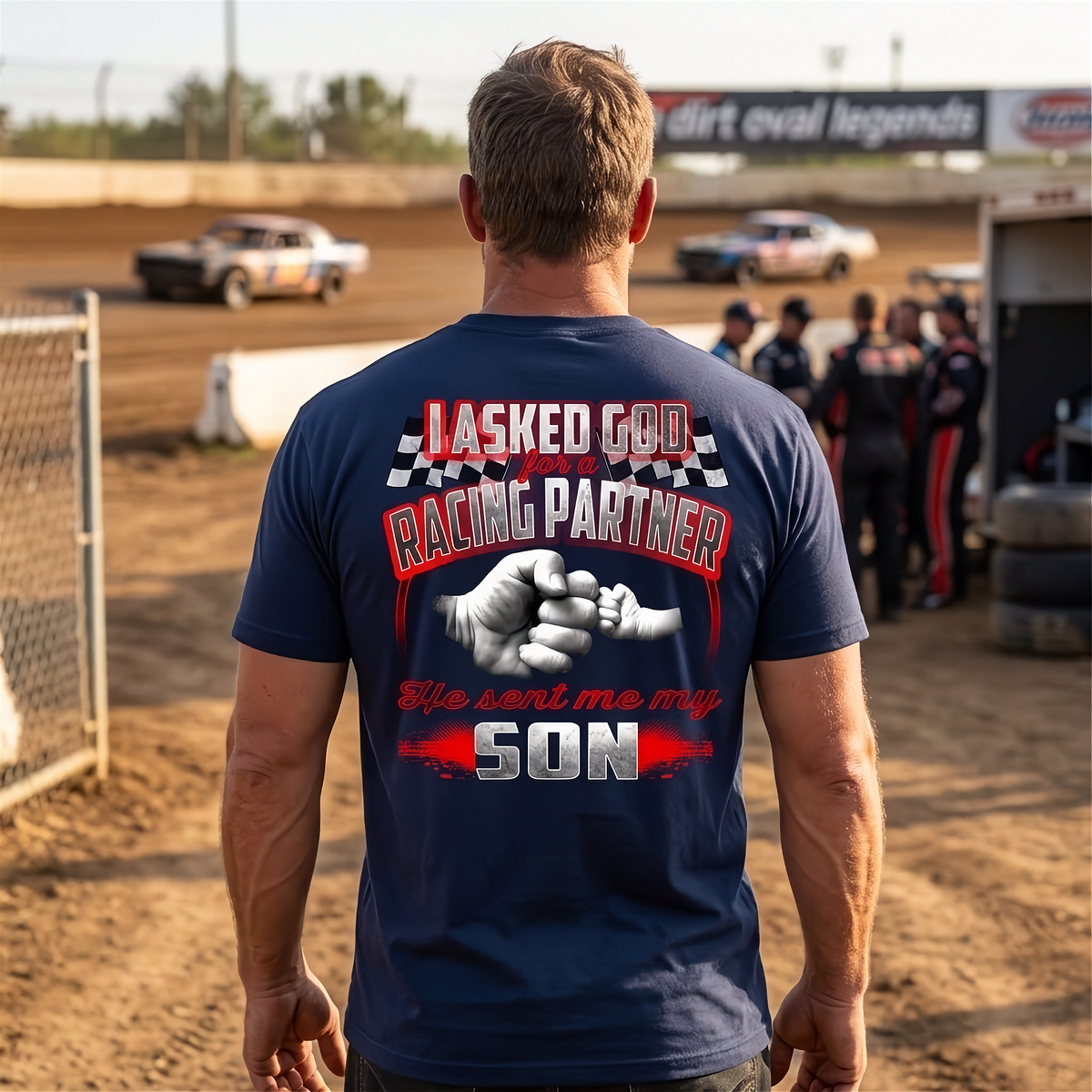 A man viewed from behind wearing a navy blue t-shirt with a large racing graphic on the back. The graphic features the text 'I ASKED GOD FOR A RACING PARTNER' and 'He sent me my SON' in red, white, and black, with a handshake illustration.