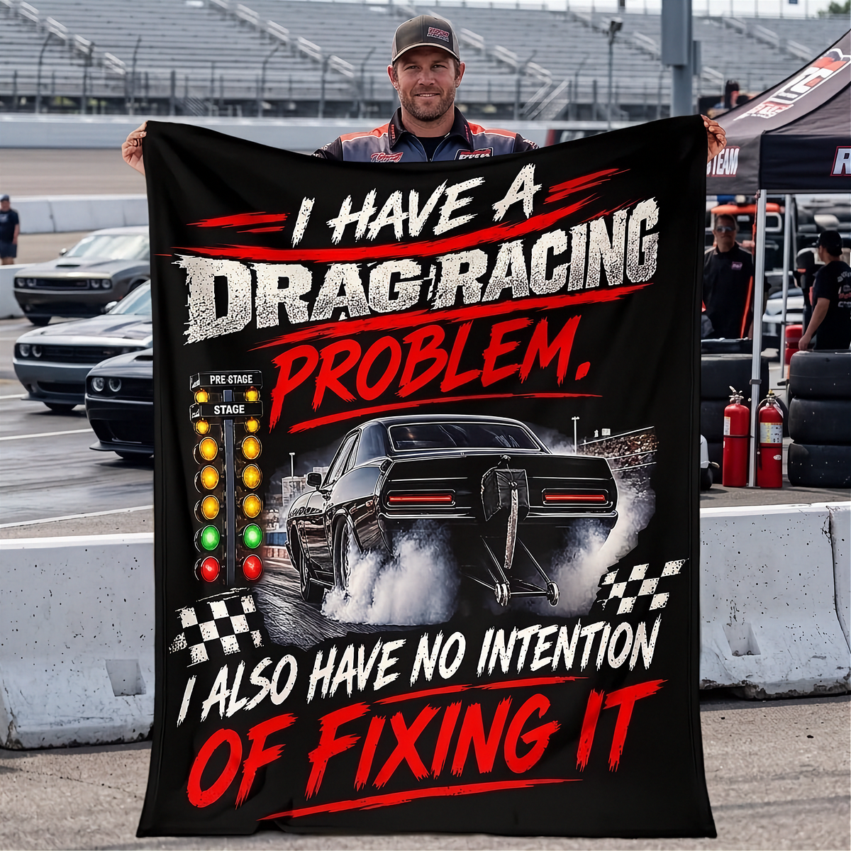 A man at a racetrack holds up a large black graphic blanket or poster featuring a drag racer car, checkered flags, and bold red and white text that reads, 'I HAVE A DRAG RACING PROBLEM. I ALSO HAVE NO INTENTION OF FIXING IT.