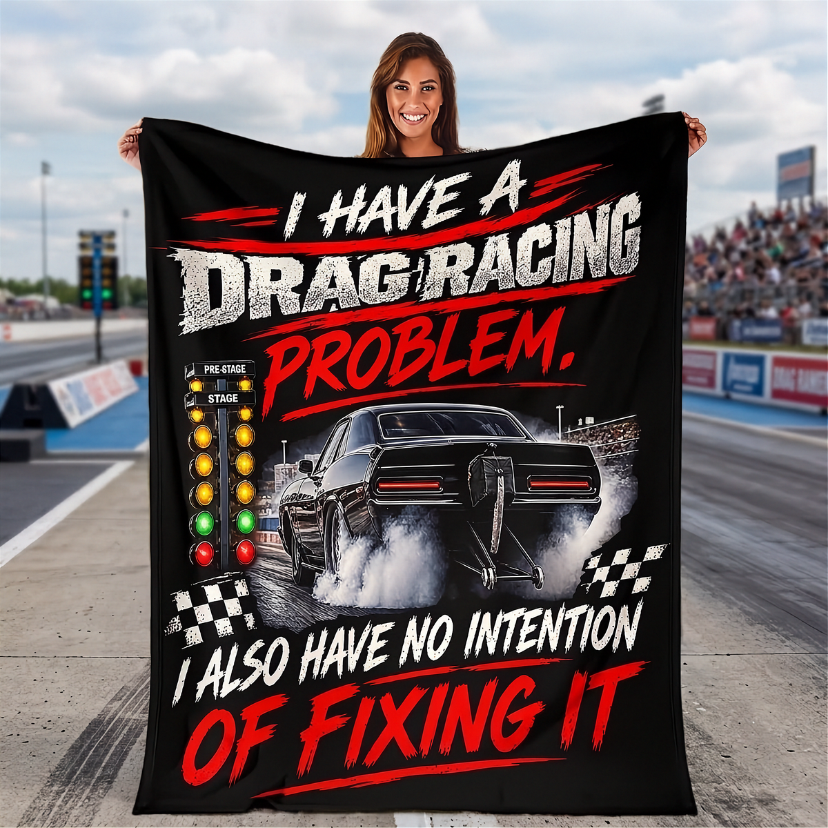A woman stands on a drag strip holding a large black printed blanket or poster that reads 'I HAVE A DRAG RACING PROBLEM. I ALSO HAVE NO INTENTION OF FIXING IT' with a car, track, and checkered flags.