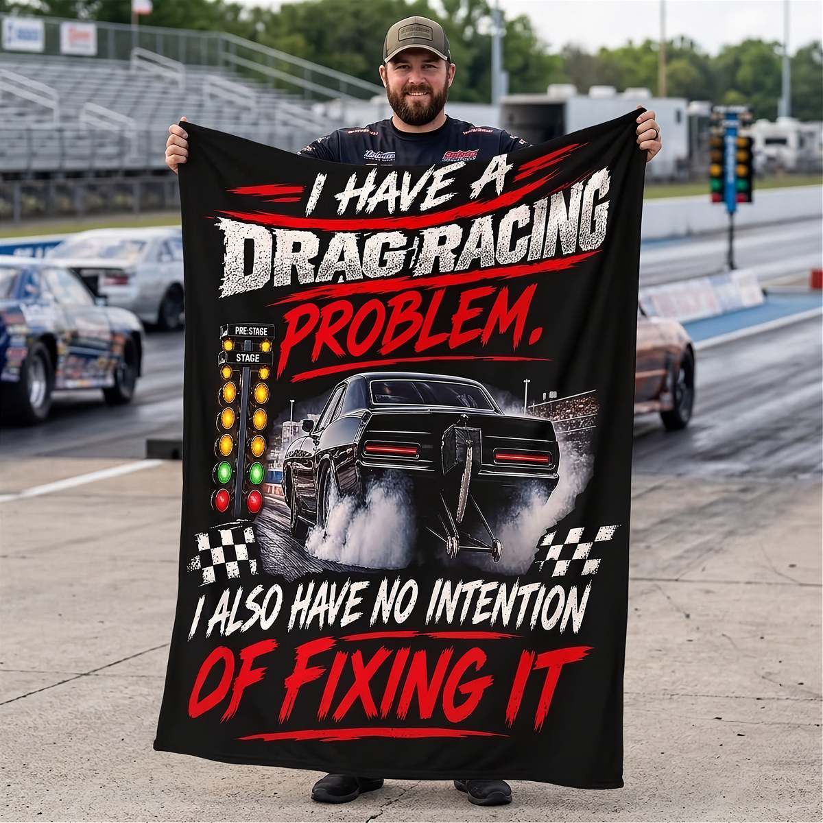 A man stands on a racetrack holding up a large black printed blanket or poster with drag racing text and a car graphic. The text reads, 'I HAVE A DRAG RACING PROBLEM, I ALSO HAVE NO INTENTION OF FIXING IT.