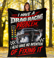 A woman stands outdoors in a wooded area holding up a large black fleece blanket or poster featuring a drag racing graphic with the text 'I HAVE A DRAG RACING PROBLEM. I ALSO HAVE NO INTENTION OF FIXING IT' and a black race car with smoke.