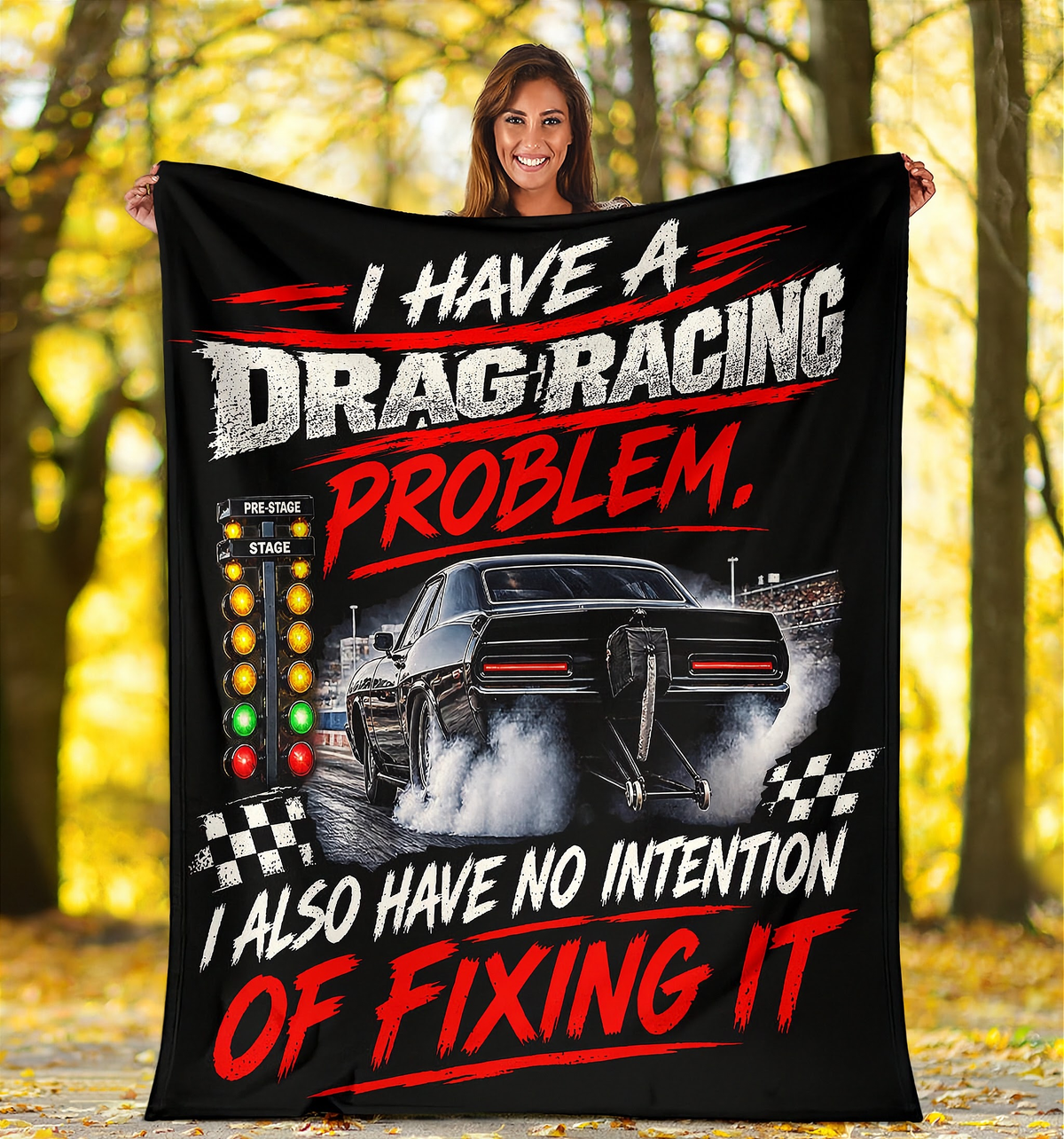 A woman stands outdoors in a wooded area holding up a large black fleece blanket or poster featuring a drag racing graphic with the text 'I HAVE A DRAG RACING PROBLEM. I ALSO HAVE NO INTENTION OF FIXING IT' and a black race car with smoke.