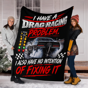 Two women holding up a large black fleece blanket featuring a drag racing graphic with the text 'I HAVE A DRAG RACING PROBLEM, I ALSO HAVE NO INTENTION OF FIXING IT' in white and red lettering.