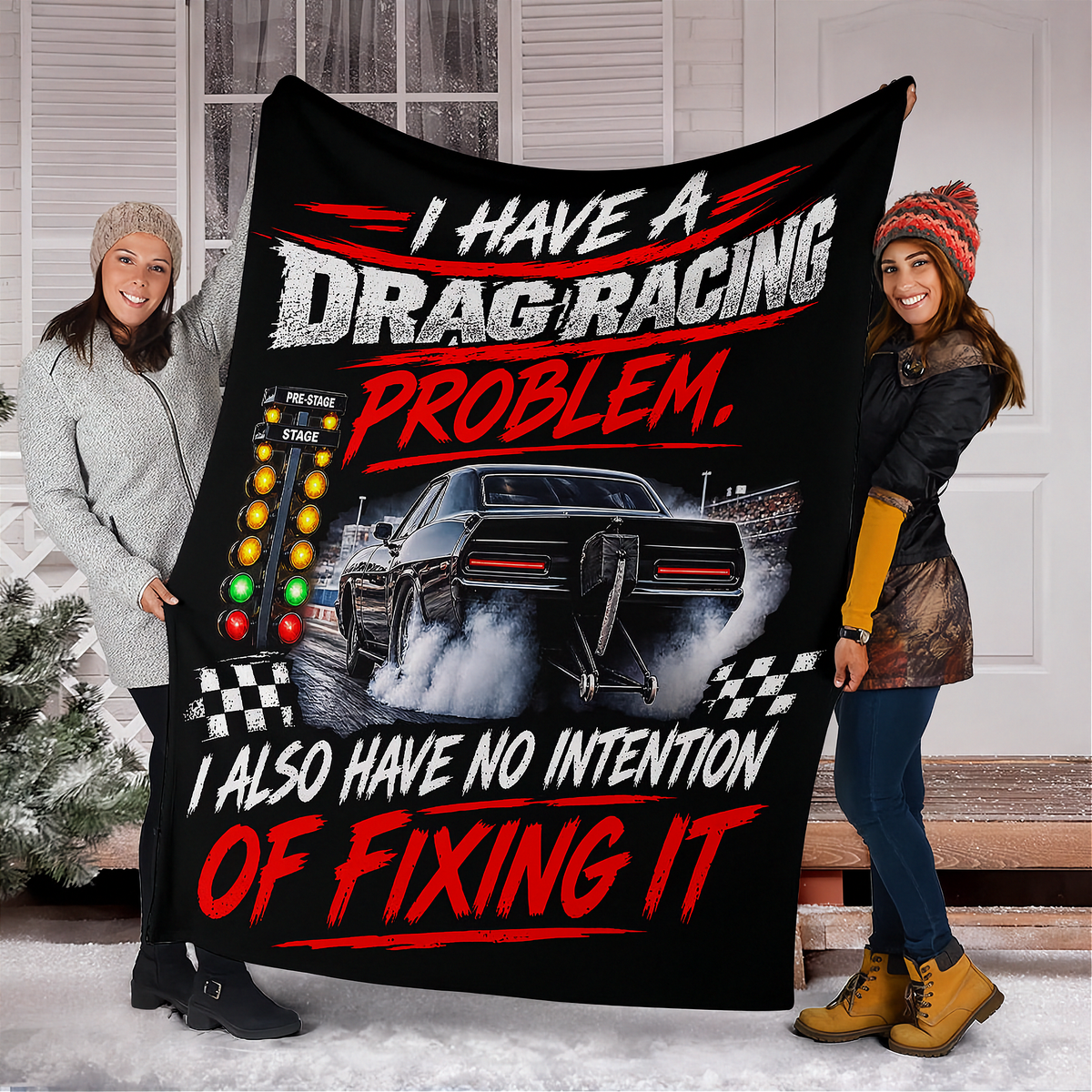 Two women holding up a large black fleece blanket featuring a drag racing graphic with the text 'I HAVE A DRAG RACING PROBLEM, I ALSO HAVE NO INTENTION OF FIXING IT' in white and red lettering.