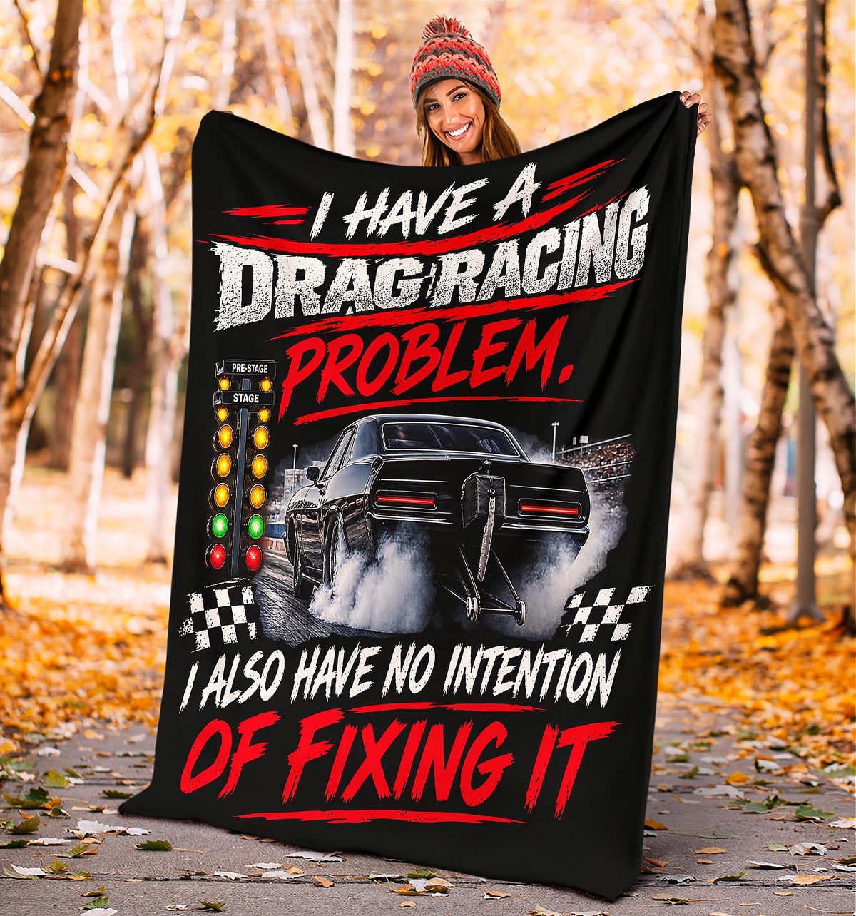 A person outdoors in a forest holding up a large black fleece blanket or throw. The blanket features a graphic of a black drag racing car with smoke, checkered flags, and bold text that reads, 'I HAVE A DRAG RACING PROBLEM, I ALSO HAVE NO INTENTION OF FIXING IT.
