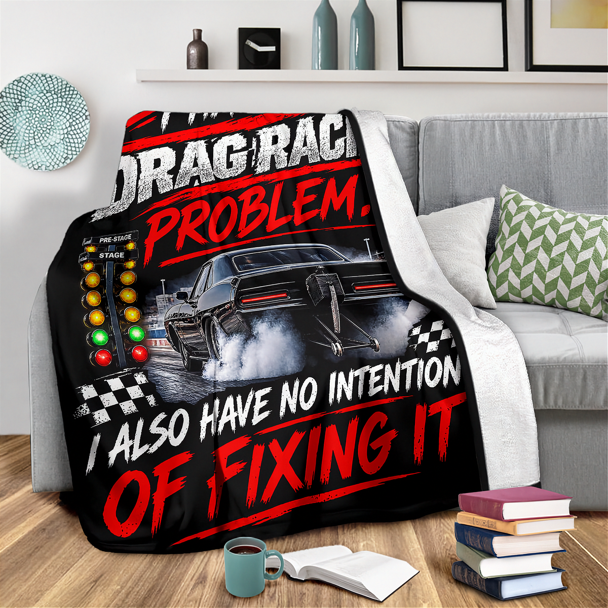 A black throw blanket draped over a gray sofa in a living room setting, featuring a graphic print of a drag racer car with the text 'DRAG RACING PROBLEM. I ALSO HAVE NO INTENTION OF FIXING IT' in white and red lettering.