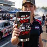 A woman at a racetrack holds up a black insulated tumbler with a clear lid. The tumbler features a graphic of a drag racing car and the text 'I HAVE A DRAG RACING PROBLEM.