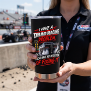 A person in a black racing media shirt holds a black insulated tumbler with a clear lid. The tumbler has a laser-engraved design featuring a drag racing car, checkered flags, and the text 'I HAVE A DRAG RACING PROBLEM, I ALSO HAVE NO INTENTION OF FIXING IT' in white and red lettering.